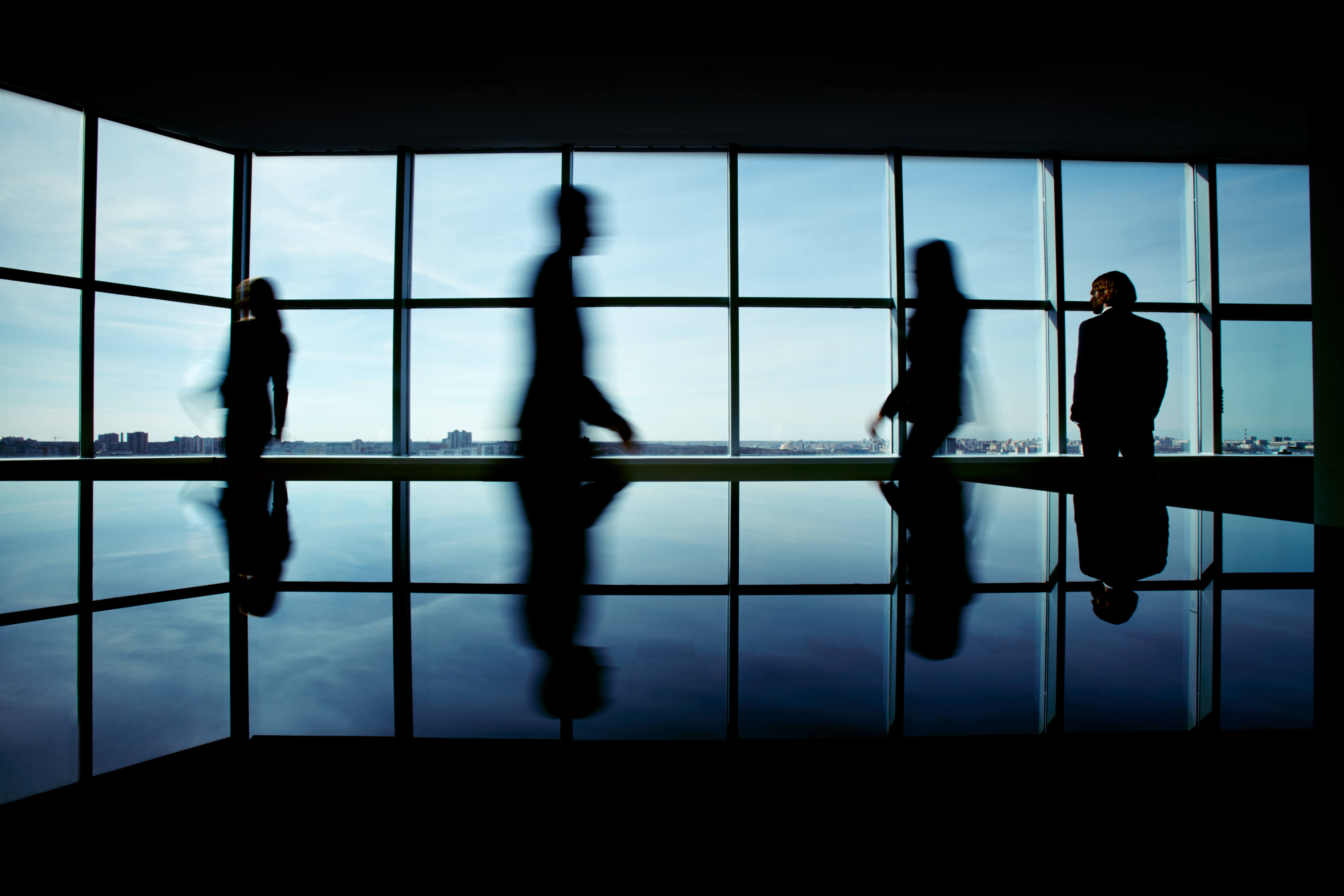 Silhouettes of business professionals walking in a modern high-rise office, reflected in a polished floor against a panoramic city skyline backdrop.