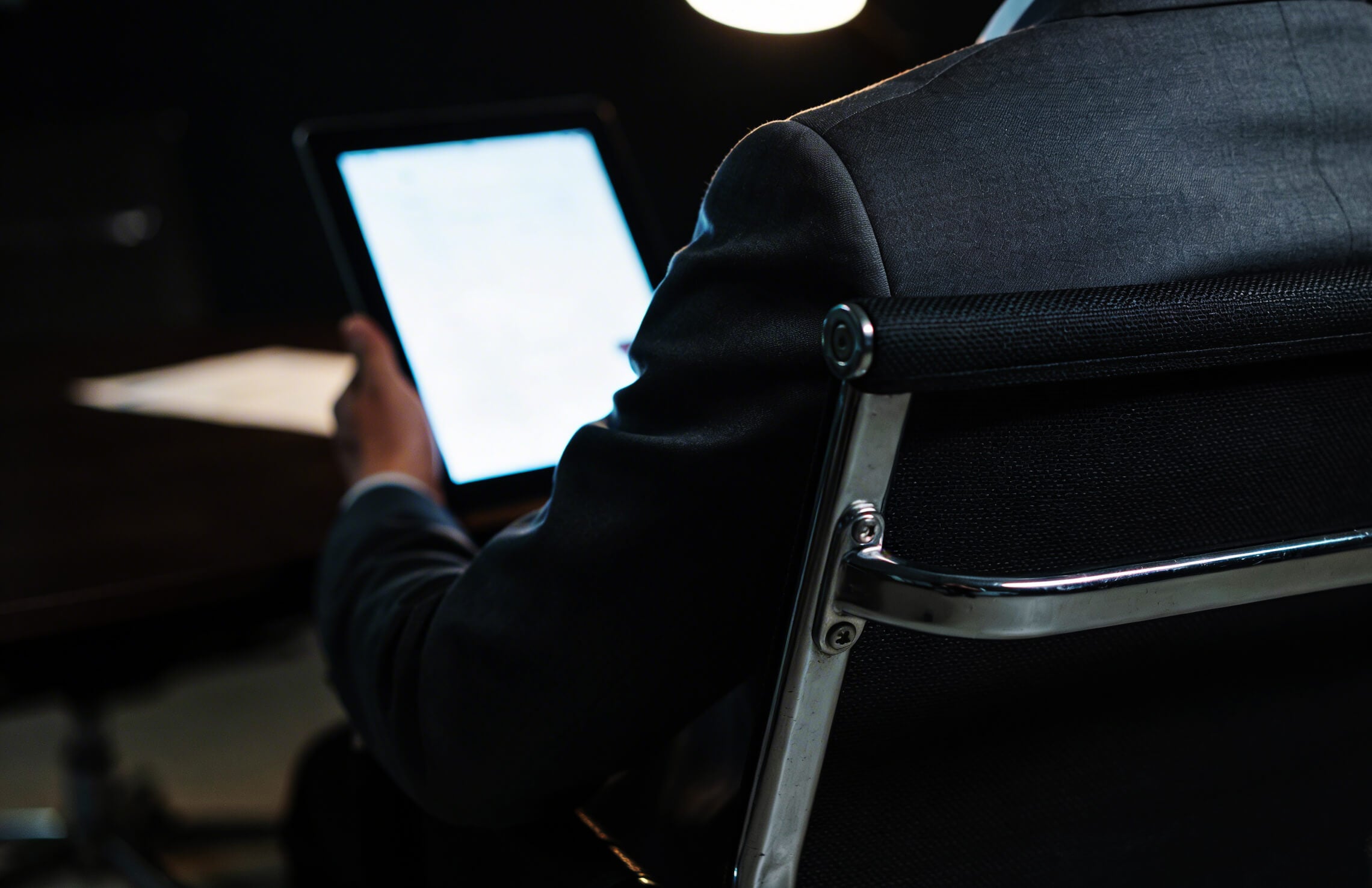 Executive in a dark suit reviewing a digital tablet in a dimly lit conference setting, viewed from behind.